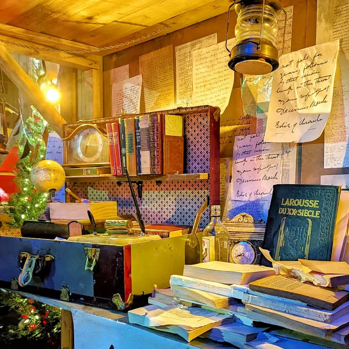 An open suitcase and stacks of books beneath a lantern, with handwritten pages covering the wall.