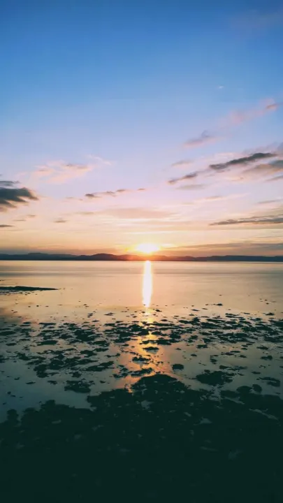 The setting sun reflects across calm water toward a rocky shoreline, with hills on the horizon.